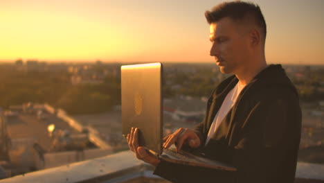 A-programmer-with-a-laptop-sits-on-the-roof-and-writes-code-at-sunset.-Remote-work-freelancer.-Freedom-to-work.-Typing-on-a-keyboard-at-sunset-with-a-view-of-the-city.